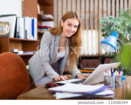 Smiling young female office employee working with laptop and papers Smiling young female office employee working with laptop and papers 119386304
