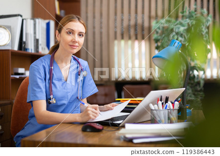 Young female general practitioner studies the patient's outpatient card, writing down the appointment Young female general practitioner studies the patient's outpatient card, writing down the appointment 119386443