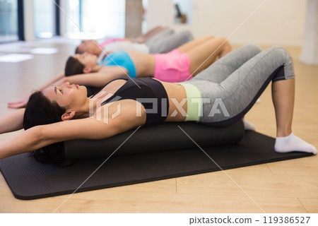 Group of people performs an exercise by placing a roll of a sports mat under their back Group of people performs an exercise by placing a roll of a sports mat under their back 119386527