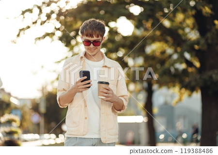 Smiling Young Man Texting on Smartphone and Holding Coffee in Morning City Street 119388486