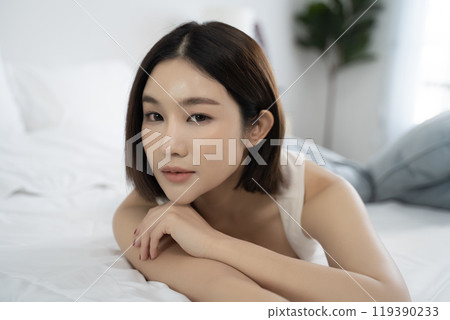 A serene close-up portrait of an Asian woman resting on a bed in a modern home, showing a peaceful expression and relaxed posture, ideal for themes of comfort, self-care, and everyday beauty. A serene close-up portrait of an Asian woman resting on a bed in a modern home, showing a peaceful expression and relaxed posture, ideal for themes of comfort, self-care, and everyday beauty. 119390233