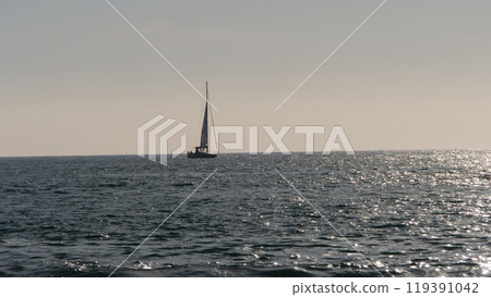 Sailboat sailing away on calm ocean at sunset with sun reflecting on water 119391042