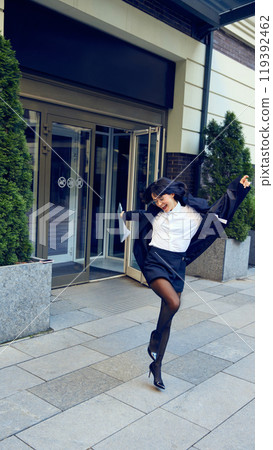 Happy young woman in stylish formal wear walking out office building with documents, getting contract for job. Employment 119392462