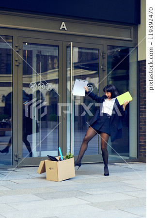Happy young woman cheerfully dancing after quitting hated job, walking out the office building with box filled with her stuffs Happy young woman cheerfully dancing after quitting hated job, walking out the office building with box filled with her stuffs 119392470
