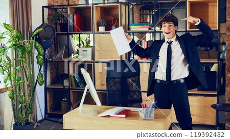 Successful deal. Happy young man, worker in formal wear standing in modern office and pointing at document, contract. New project 119392482
