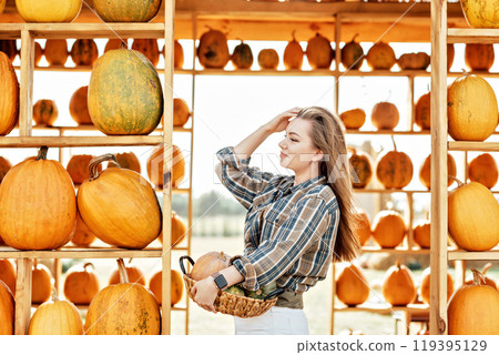young woman stands at pumpkin festival, holding large orange and green pumpkin. She is surrounded by rows of pumpkins and a wooden display. The autumn farm setting captures the harvest season vibe. 119395129