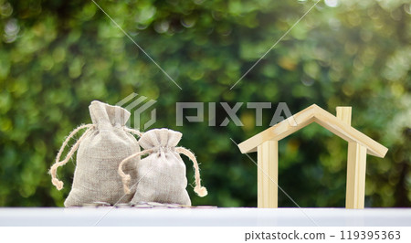 Brown burlap sacks filled with coins and some scattered on a white surface and wooden house model placed nearby, with blurred background of bushes, growth in savings and financial planning. 119395363