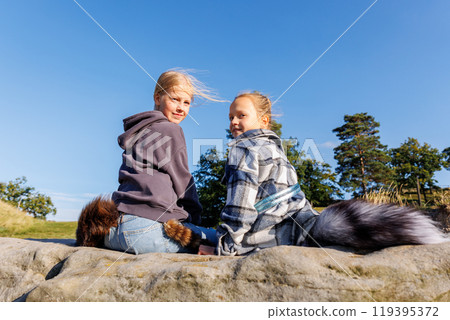 Back view two cute little girls friends wearing casual hoodies plaid jackets and attached fox fur tails enjoy sit on rock on warm sunny day outdoors. Clear blue sky trees woods background Back view two cute little girls friends wearing casual hoodies plaid jackets and attached fox fur tails enjoy sit on rock on warm sunny day outdoors. Clear blue sky trees woods background 119395372