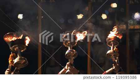 Varanasi, India. Man makes movements with a candle holder with lit candles on the Ganga Maha Aarti ceremony begins. Brahman priest wearing red and yellow dress doing traditional ganga aarti at ghat Varanasi, India. Man makes movements with a candle holder with lit candles on the Ganga Maha Aarti ceremony begins. Brahman priest wearing red and yellow dress doing traditional ganga aarti at ghat 119395386