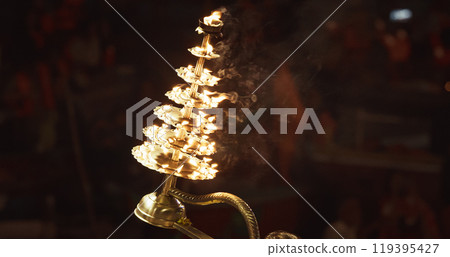 Varanasi, India. Man makes movements with a candle holder with lit candles on the Ganga Maha Aarti ceremony begins. Brahman priest wearing red and yellow dress doing traditional ganga aarti at ghat Varanasi, India. Man makes movements with a candle holder with lit candles on the Ganga Maha Aarti ceremony begins. Brahman priest wearing red and yellow dress doing traditional ganga aarti at ghat 119395427