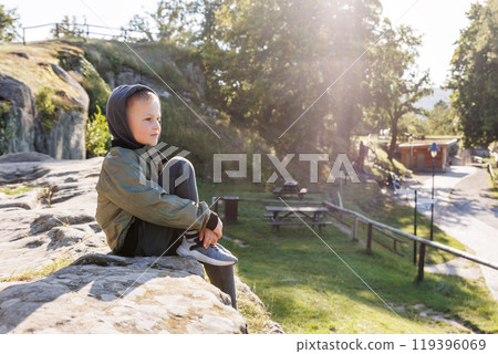 Little cute adorable thoughtful kid boy in hoodie and jacket sitting on rock enjoy dreaming in warm sunlight at city nature park. Pensive happy child  portrait outdoors. Carefree childhood concept 119396069