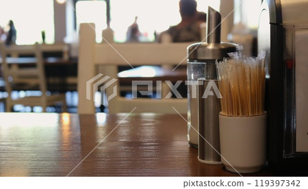 toothpicks, salt shaker and pepper shaker standing on empty wooden table on the right and blurred background of cafe, restaurant or cafeteria with couple silhouettes eating 119397342