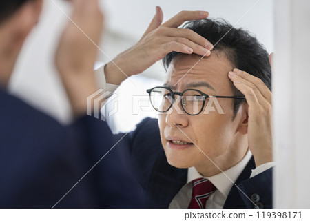 A businessman in his late 30s who is concerned about his hairline while looking in the mirror A businessman in his late 30s who is concerned about his hairline while looking in the mirror 119398171