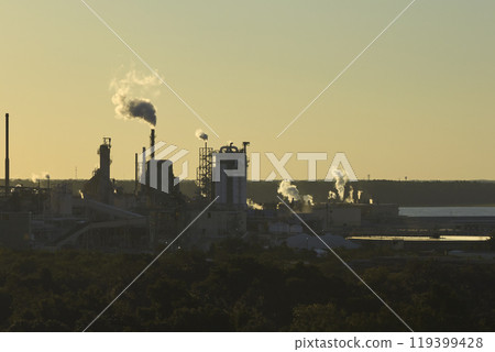Large factory structure with smoke from production process rising up polluting atmosphere at manufacturing yard. Industrial site at sunset 119399428