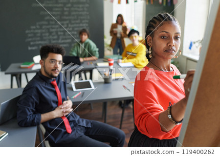 Black young woman using flip chart during seminar in school classroom with students and teacher watching, copy space 119400262