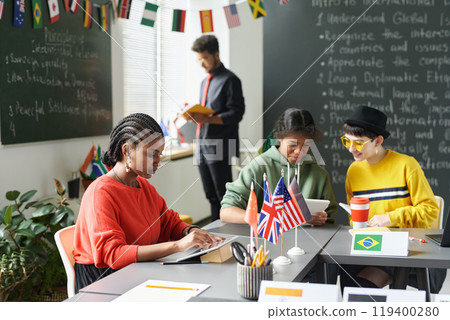 Diverse group of students sitting at table during lesson in school classroom and using gadgets, copy space 119400280
