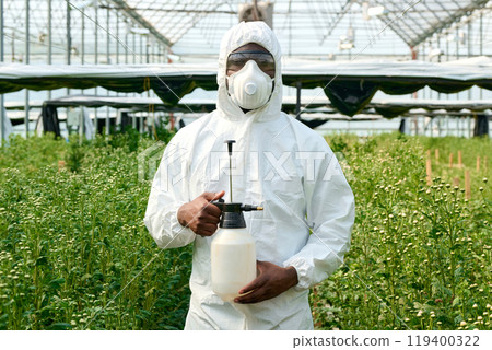 Waist up portrait of man wearing protective suit in greenhouse and holding spray bottle while looking at camera 119400322