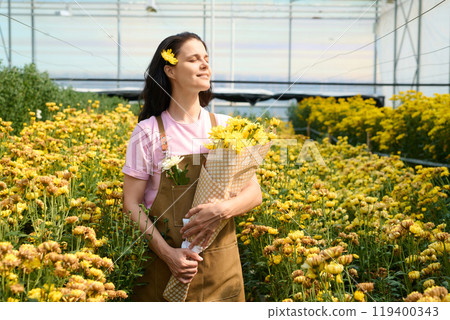 Portrait of smiling young woman enjoying sunlight with eyes closed while holding bouquet of yellow daisies in flower greenhouse, copy space 119400343