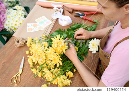 Close up of young woman arranging flower composition with yellow flowers on wooden table with tools and craft paper in flower shop copy space Close up of young woman arranging flower composition with yellow flowers on wooden table with tools and craft paper in flower shop copy space 119400354