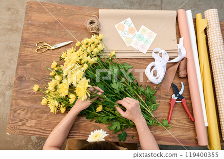 Top view of female hands arranging flower composition on wooden table with tools and craft paper in flower shop copy space Top view of female hands arranging flower composition on wooden table with tools and craft paper in flower shop copy space 119400355