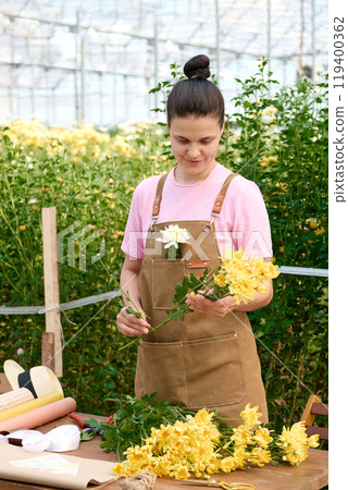 Vertical portrait of young woman arranging bouquet of dainty yellow flowers at market sale in sunlit plantation 119400362