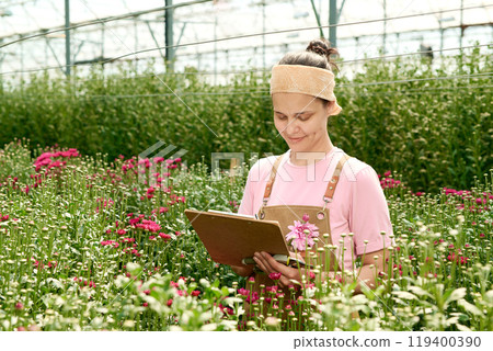 Waist up portrait of smiling Caucasian young woman holding clipboard and taking notes standing in industrial greenhouse plantation lit by sunlight copy space 119400390