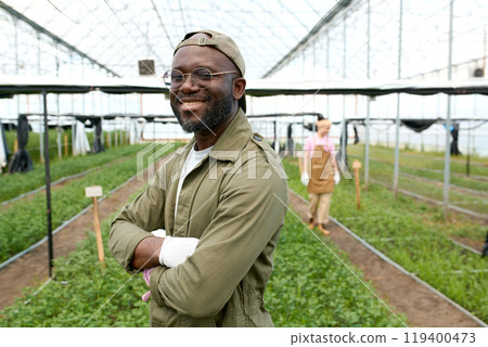 Waist up portrait of smiling African American man wearing glasses and working as gardener posing confidently standing with arms crossed in glass greenhouse copy space 119400473