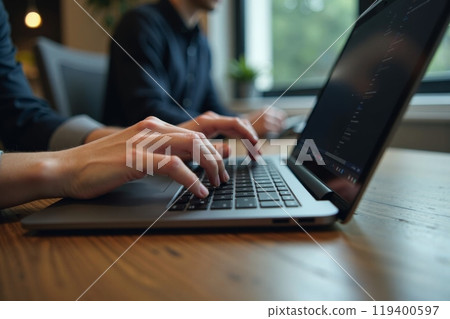 Close-up of hands typing on laptop keyboard in modern office setting with window view. Focus on hands, while background features a blurred image of another professional working 119400597