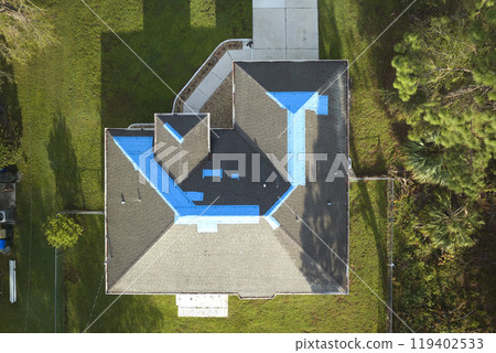 Aerial view of damaged in hurricane Ian house roof covered with blue protective tarp against rain water leaking until replacement of asphalt shingles Aerial view of damaged in hurricane Ian house roof covered with blue protective tarp against rain water leaking until replacement of asphalt shingles 119402533