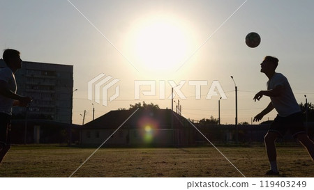 Silhouette of professional footballers kicking soccer ball with their heads on stadium at sunset. Two sportsmen showing tricks with ball while passing it to each other at field. Freestyle football 119403249