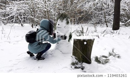 Happy woman building snowman at winter forest. Young girl having active leisure at snowy park. Cheerful female having fun outdoor. Concept of a winter vacation. 119403261