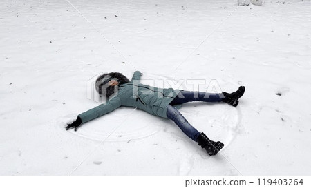 Happy woman lying on snowy lawn at forest and making angel. Young girl playing in snow and enjoying wintertime. Cheerful lady having fun outdoor. Concept of a winter vacation. 119403264