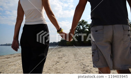 Couple holding hands of each other and walking at coast. Happy pair going along seashore at sunny day. Lovers spending time together at resort. Concept of vacation or holiday. Slow mo Couple holding hands of each other and walking at coast. Happy pair going along seashore at sunny day. Lovers spending time together at resort. Concept of vacation or holiday. Slow mo 119403945