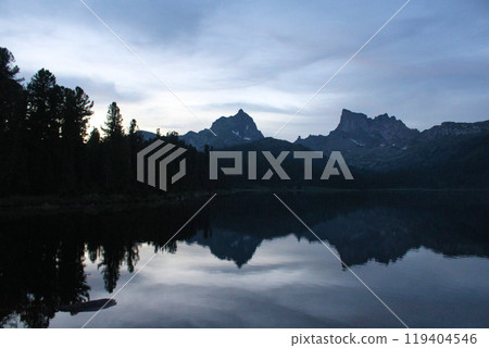 Silhouettes of two rocky mountain peaks and pine trees are reflected in the water of the lake in twilight 119404546