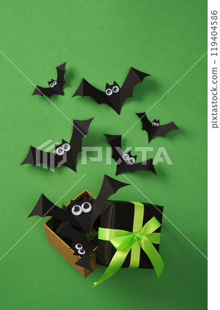 Halloween gift box with green neon ribbon with paper bats flying out of it. Green background. 119404586