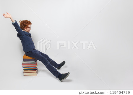 Funny redhead child boy in school uniform sitting on stack of books with hands up  119404759