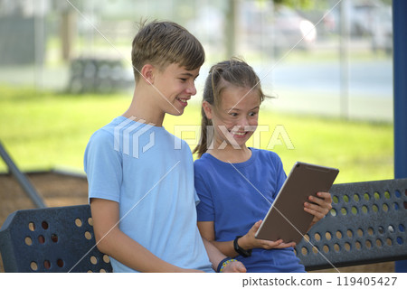 Two happy children, girl and boy looking at screen of digital tablet, reading, studying or gaming sitting on bench outdoors on summer sunny day 119405427