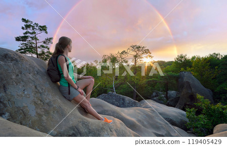 Sportive woman sitting alone taking a break on hillside trail. Female hiker enjoying view of evening nature from rocky cliff on wilderness path. Active lifestyle concept Sportive woman sitting alone taking a break on hillside trail. Female hiker enjoying view of evening nature from rocky cliff on wilderness path. Active lifestyle concept 119405429