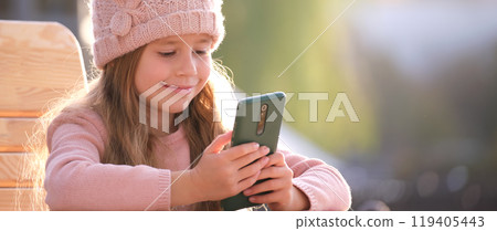 Portrait of cute little child girl in pink hat sitting alone at street cafe playing game on sellphone 119405443