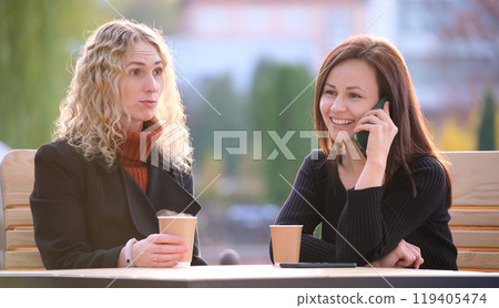 Female friends spending time at street cafe outdoors while one girl is talking happily on mobile phone and another waiting frustrated at offensive behaviour of her mate. Friendship problems concept Female friends spending time at street cafe outdoors while one girl is talking happily on mobile phone and another waiting frustrated at offensive behaviour of her mate. Friendship problems concept 119405474