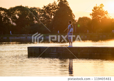 Back view of lonely woman standing on lake side on warm evening. Solitude and relaxation concept. Back view of lonely woman standing on lake side on warm evening. Solitude and relaxation concept. 119405481