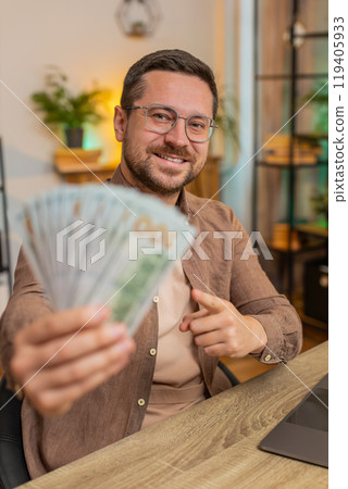 Young businessman freelancer holding fan of cash and showing thumbs up sitting at home office table 119405933