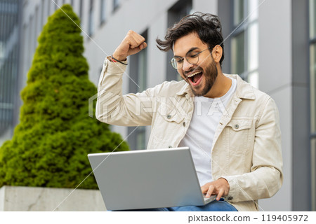 Happy Indian man working on laptop celebrate success win money get online good news outdoors street 119405972