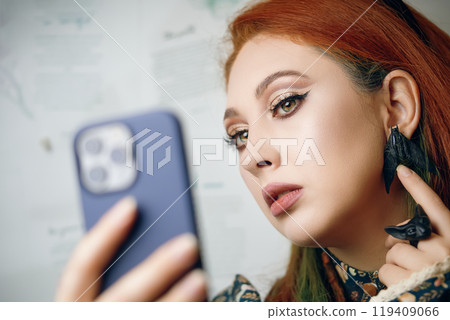 Red-haired woman takes selfie on front camera of modern smartphone. Female blogger films herself for fashion and makeup blog. Cute girl points her finger at her butterfly earrings. Gothic aesthetic. Red-haired woman takes selfie on front camera of modern smartphone. Female blogger films herself for fashion and makeup blog. Cute girl points her finger at her butterfly earrings. Gothic aesthetic. 119409066
