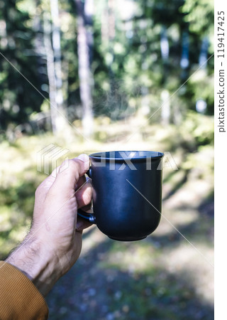 Hand holding a cup of drink in outdoor camp. Hot coffee, tea or mulled wine in a camp in nature in the forest. Travel, nature, recreation 119417425