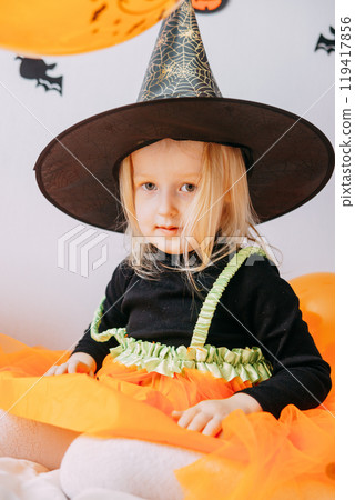 Children's Halloween - a girl in a witch hat and a carnival costume with airy orange and black balloons at home. Ready to celebrate Halloween 119417856