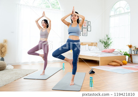Couple Asian women exercise together with yoga posture by stand and hands together over head also stay on mat in room with day light. 119421810