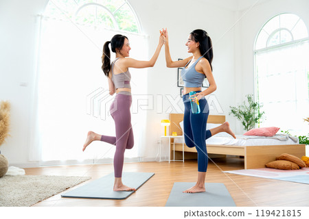 Asian women relax after yoga exercise by stand with hold water bottle and touch their hands together also raise legs up in room with day light. 119421815
