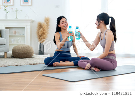 Couple Asian women with exercise clothes sit on yoga mat and hold bottle of water then cheers together and relax after workout in the house. 119421831