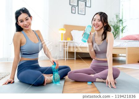Asian women with exercise clothes sit on yoga mat and hold bottle of water also look at camera with relax after workout in the house. Asian women with exercise clothes sit on yoga mat and hold bottle of water also look at camera with relax after workout in the house. 119421836